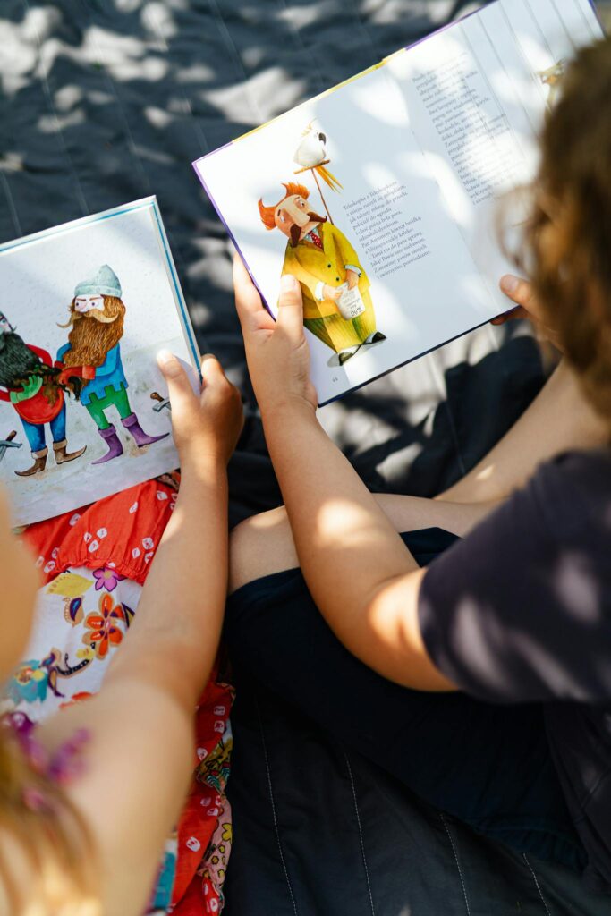 Two children enjoying storybooks in a sunny outdoor setting with leaf shadows.