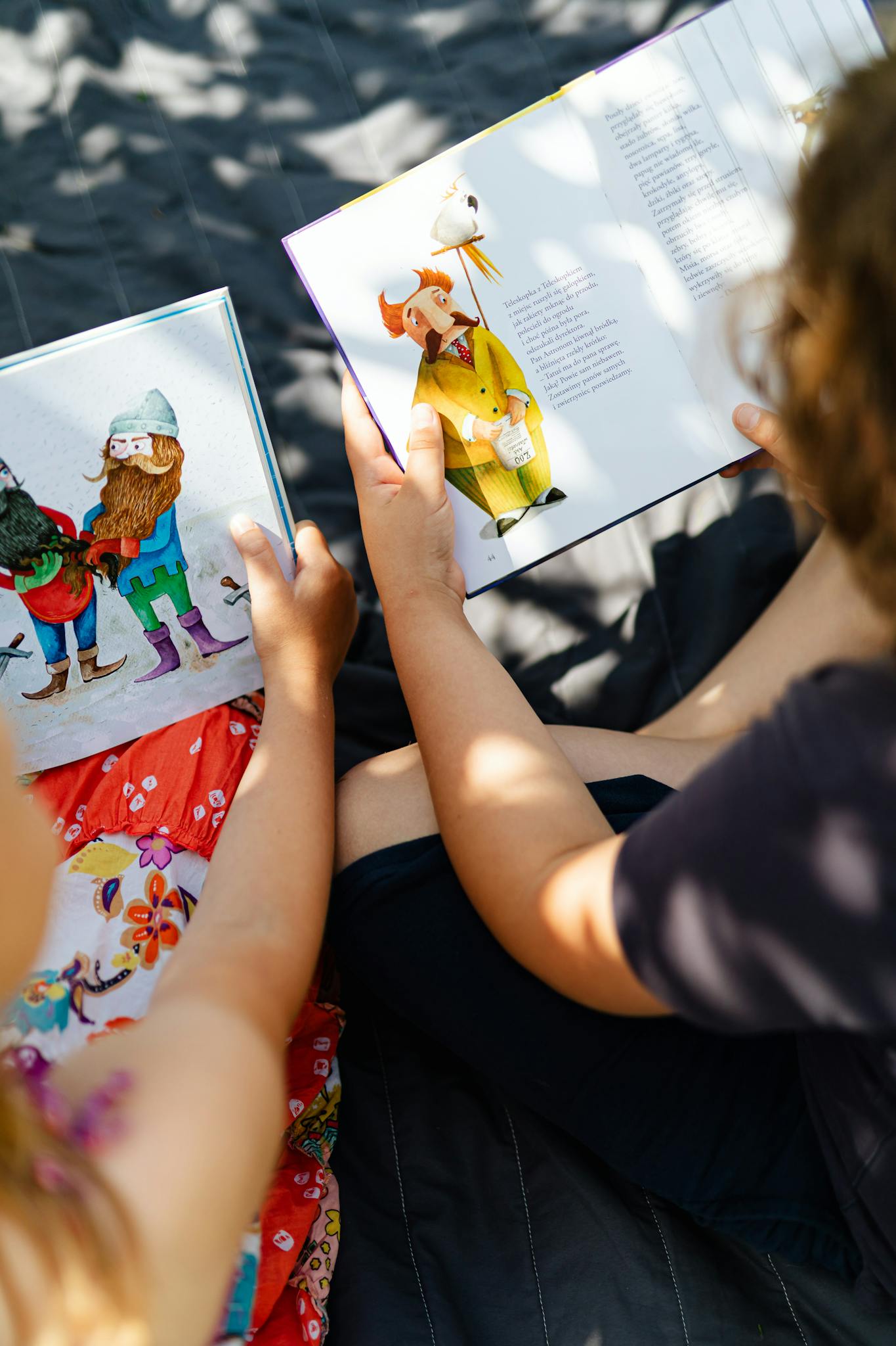 Two children enjoying storybooks in a sunny outdoor setting with leaf shadows.