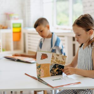 Two children reading books at a bright indoor classroom table filled with colorful supplies.