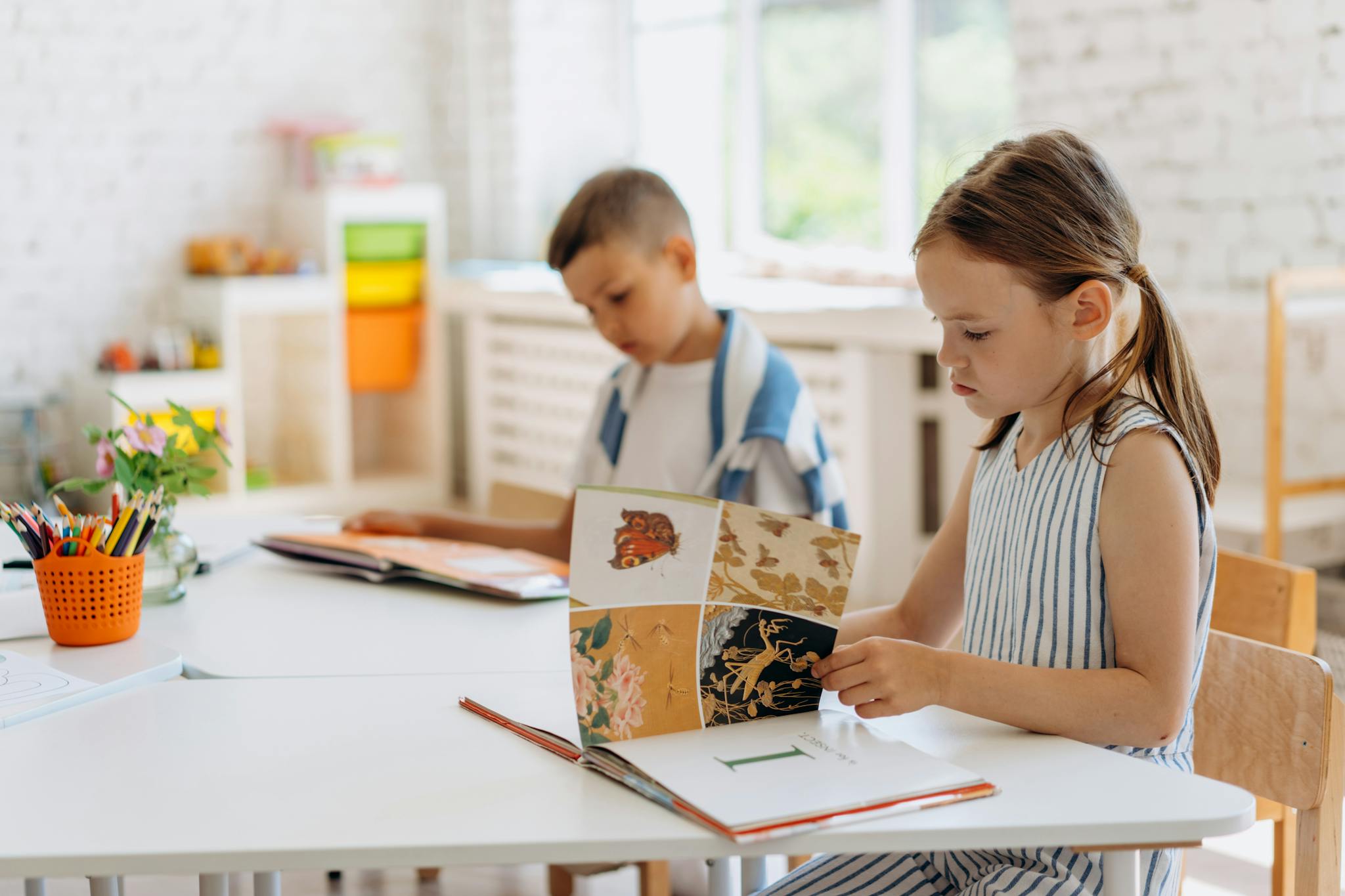 Two children reading books at a bright indoor classroom table filled with colorful supplies.