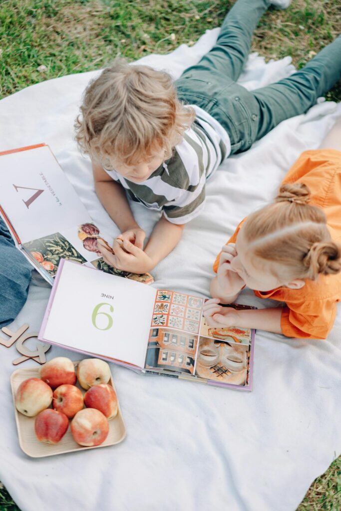 Two children reading books on a picnic blanket with apples outdoors. Perfect for educational or lifestyle themes.